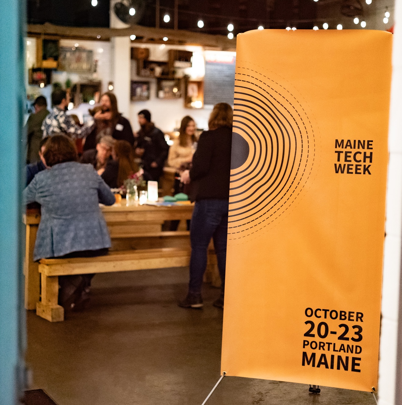 A Maine Tech Week banner in the doorway of a room full of people at long tables